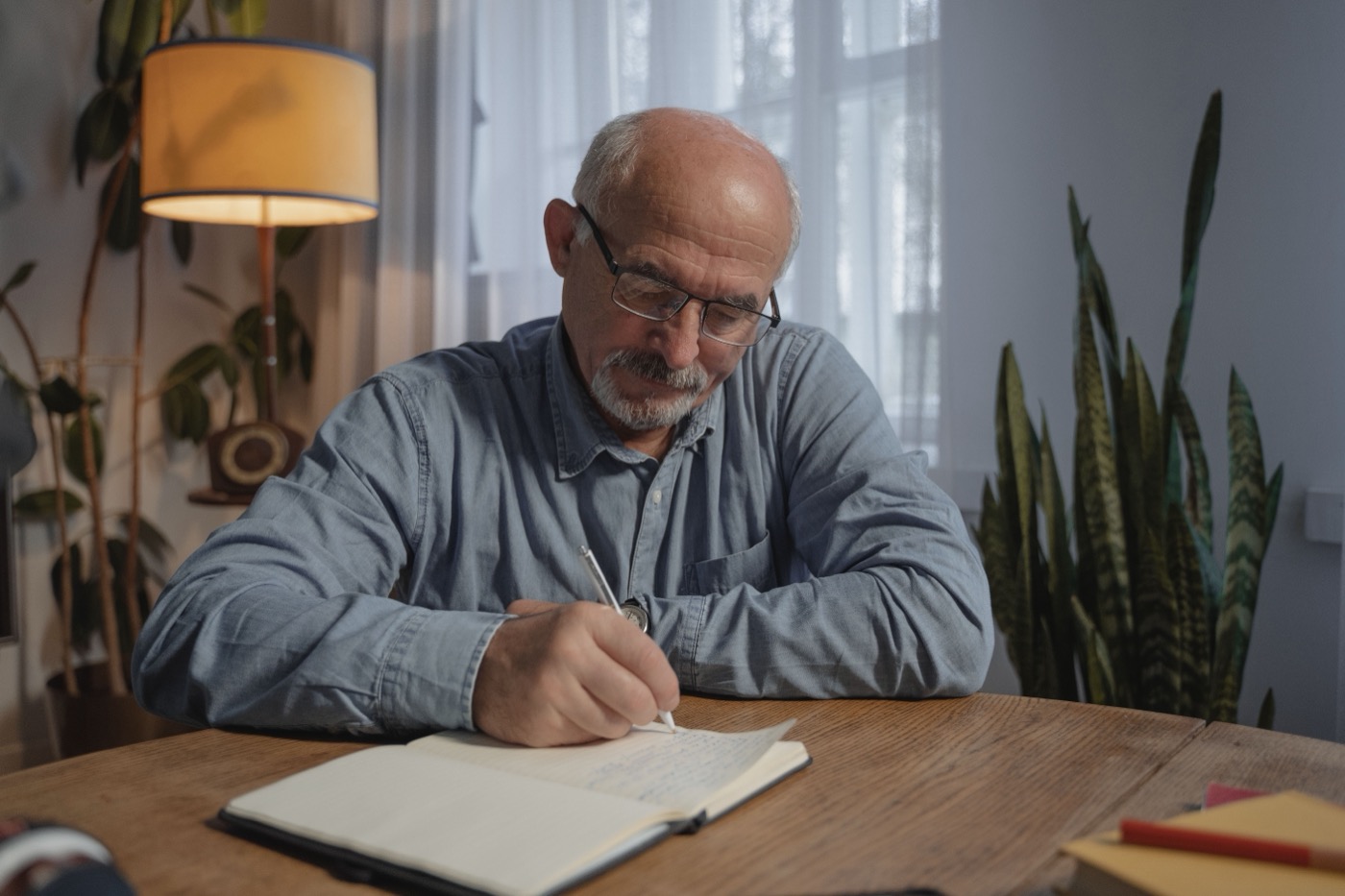 Ein älterer Mann mit Brille schreibt in ein Notizbuch an seinem Schreibtisch